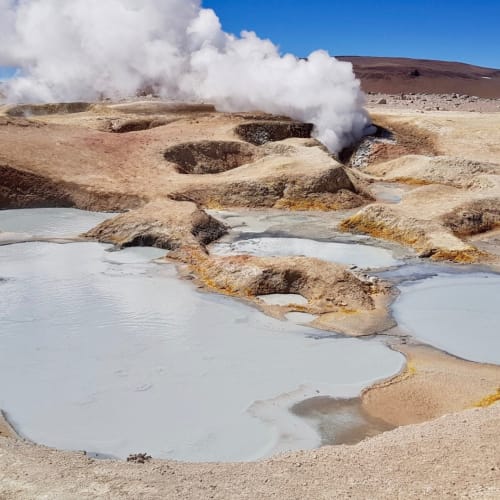 Geysers and thermal pools in the Chilean Atacama Desert with steam rising into a clear blue sky.