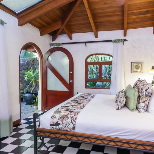 Cozy bedroom with a wooden ceiling, patterned bed linen, a checkered black and white floor, green accent wall, and typical arched windows and an arched door leading to an outdoor stone patio with plants.