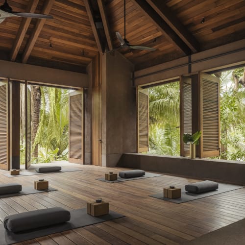 Innenraum eines offenen Yoga-Studios mit Holzboden, Holzwänden und großen offenen Fenstern mit Blick auf grüne Palmen und tropische Vegetation.