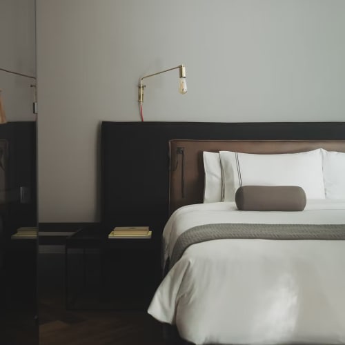 Modernes Schlafzimmer mit großem Bett, weißer Bettwäsche, grauer Decke, zylindrischen Kissen, einem schwarzen Nachttisch mit Büchern und einer messingfarbenen wandmontierten Leselampe.
