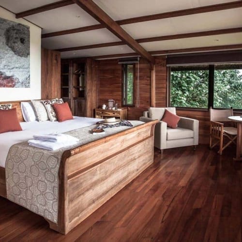 A spacious bedroom in a wooden lodge with a large bed, armchair, round table, and large windows showing greenery outside.