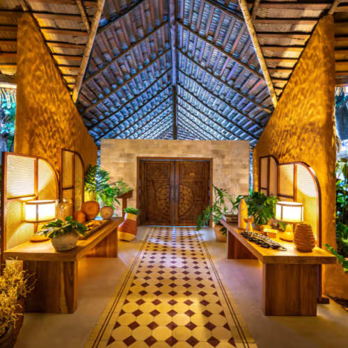 Innenbereich eines tropischen Öko-Resort-Restaurants mit Holztischen und dekorativen Pflanzen unter einer hohen Strohdach-Decke.