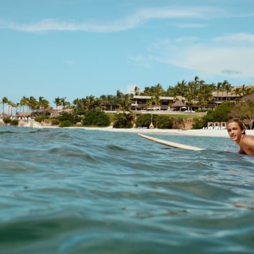 Eine Person liegt auf einem Surfbrett und paddelt im Meer in der Nähe eines tropischen Strandresorts mit Palmen unter einem leicht bewölkten Himmel.