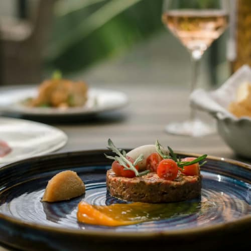 Ein Gourmetgericht mit einer geschichteten Tartare, garniert mit halbierten Kirschtomaten und Kräutern, serviert auf einem dunklen Keramikteller mit einer orangenen Sauce und einem kleinen Brotzstück, auf einem Tisch mit unscharfem Hintergrund mit einem Glas Weißwein und einer Schale mit Brot.