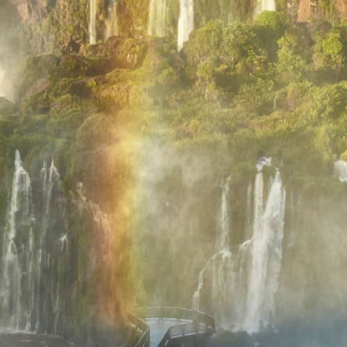 Mehrere Wasserfälle stürzen über grüne Klippen mit einem Regenbogen in der Nähe des Bodens, und ein Spazierweg erstreckt sich über das Wasser im Vordergrund.