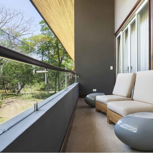 A cozy balcony with two beige armchairs and two glossy gray side tables, overlooking lush green trees through glass sliding doors.