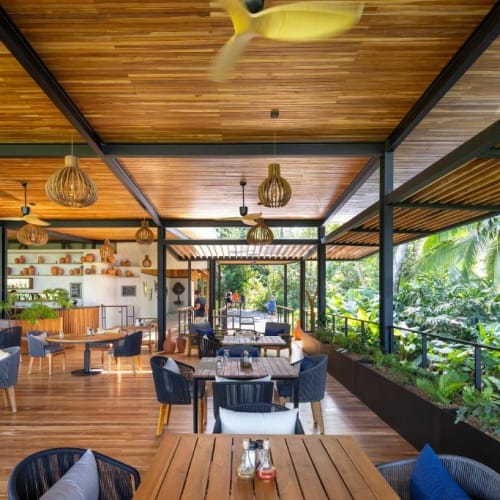 Freiluftrestaurant mit Holzboden und Holzbalkendecke, modernen Korbmöbeln, umgeben von üppigem tropischem Grün.
