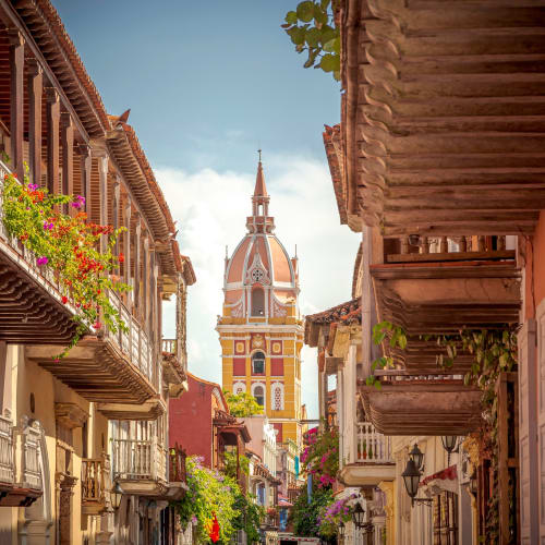 Colorful colonial-style buildings with balconies and flowers line a narrow street, with a tall historic tower in the background.