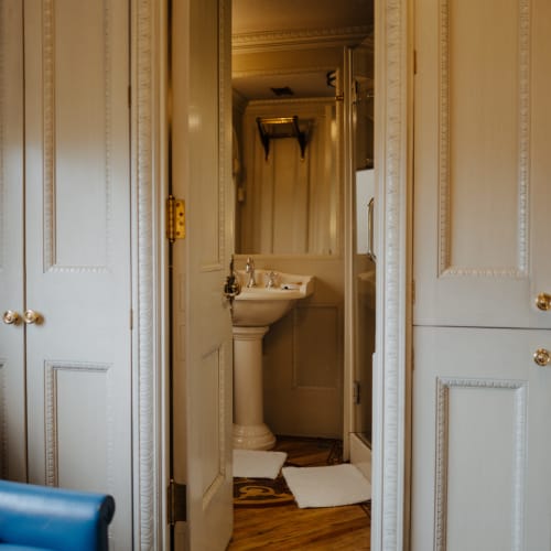 Blick durch eine halb geöffnete Tür, die ein Badezimmer mit einem Standwaschbecken, weißen Badematten und Holzfußboden in einem Vintage-Stil zeigt.