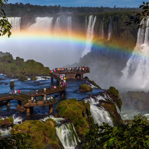 Eine Aussichtsplattform mit Touristen blickt auf die mächtigen Wasserfälle von Iguazu, mit einem Regenbogen, der über den Nebel spannt.