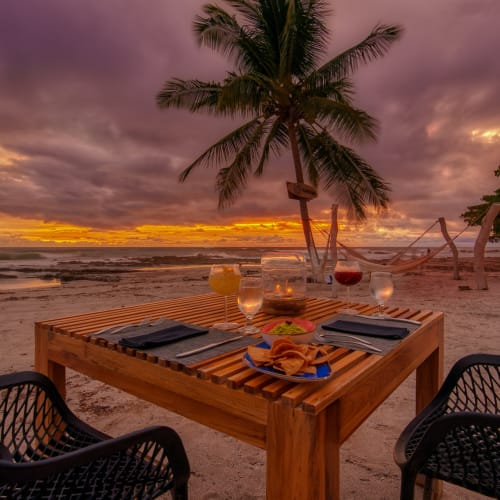 A wooden table set for dinner on a sandy beach at sunset, with palm trees in the background and two black chairs around the table.