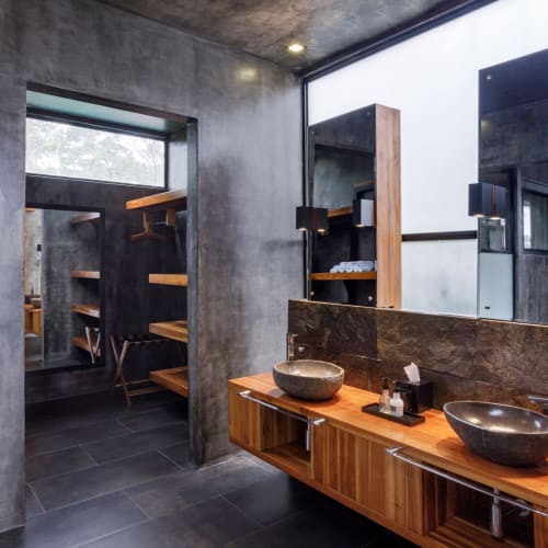 Modernes Badezimmer mit rustikalen Betonwänden, zwei steinernen Aufsatzwaschbecken auf einer Holzarbeitsplatte, großen Spiegeln und schwarzen Wandlampen, angrenzend an einen begehbaren Kleiderschrank mit Holzregalen.