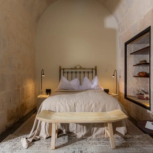 Gemütliches Schlafzimmer mit Steinwänden, einem vintage Metallbett mit beiger Bettwäsche, Nachttischlampen, einer Holzbank am Fußende des Betts und einem kleinen runden Tisch mit Lampe und Buch.