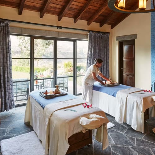 Eine Frau bereitet Massagetische in einem gemütlichen Spa-Raum mit großen Fenstern, Holzbalken und Steinboden vor.