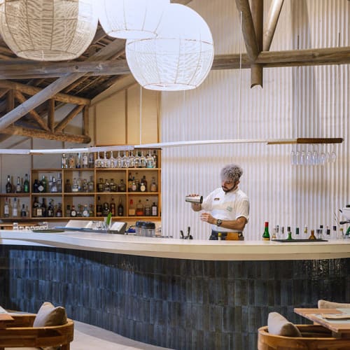 Barkeeper bereitet einen Cocktail hinter einer modernen Bar mit weißen runden Hängeleuchten und Holzmöbeln in einem gemütlichen Restaurant vor.