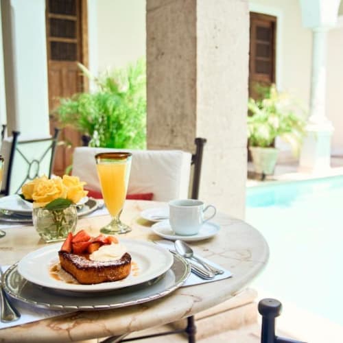 Frühstücksarrangement auf einem runden Marmortisch mit French Toast, garniert mit Erdbeeren und Sahne, einer Kaffeetasse, zwei Gläsern Orangensaft und einer Vase mit gelben Rosen, neben einem Pool in einem hellen, offenen Terrassenbereich.