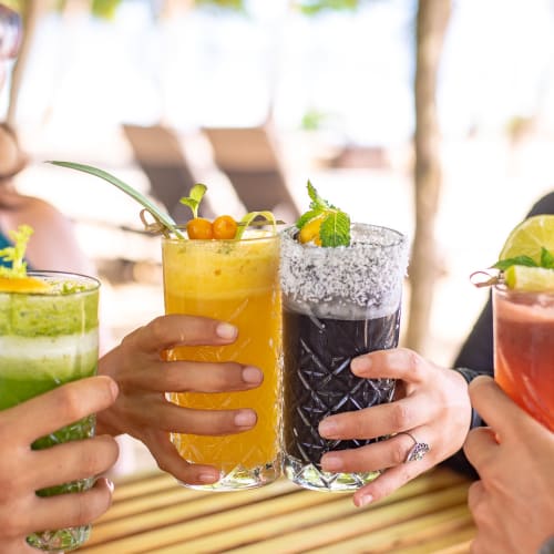Drei Frauen, die draußen an einem Strandresort mit Bäumen im Hintergrund bunte Cocktails genießen.