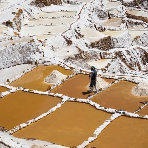 A person walks on salt evaporation ponds with a mountainous landscape in the background.