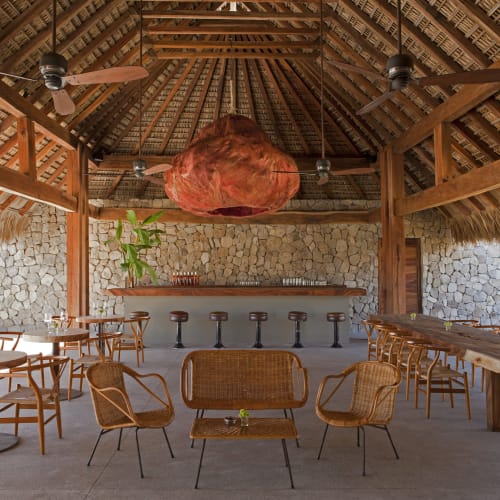 Freiluftbar mit Strohdach, Steinmauern, Holztischen und Stühlen sowie einer Bartheke mit Barhockern unter Deckenventilatoren und einem großen orangefarbenen hängenden Dekorationselement.