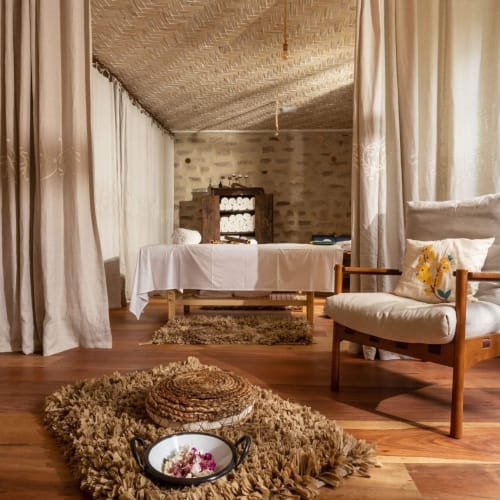 A cozy spa room with a wooden floor, a comfortable cushioned chair, a massage table covered with a white sheet, and natural light coming through a window with green frames.