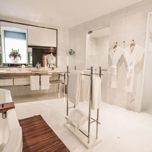 Ein modernes, helles Badezimmer mit einem großen Spiegel über einem Doppelwaschbecken, einer weißen Badewanne mit Holzablage, zwei weißen Bademänteln an der Wand und einem Handtuchhalter in der Raummitte.