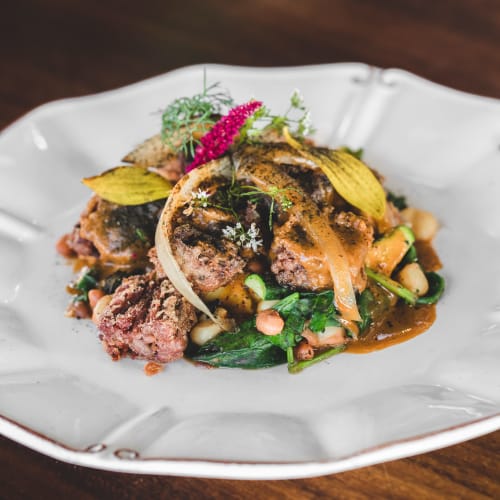 Ein Teller mexikanisches Essen mit gewürztem gegrilltem Fleisch, Gemüse, Bohnen und garniert mit Kräutern und essbaren Blumen auf einem weißen Keramikteller.