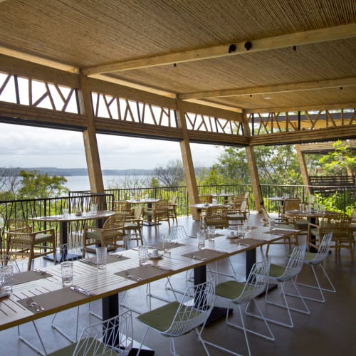 Bright, modern dining area with large windows overlooking a lush landscape and water, featuring wooden beams and assorted tables and chairs.