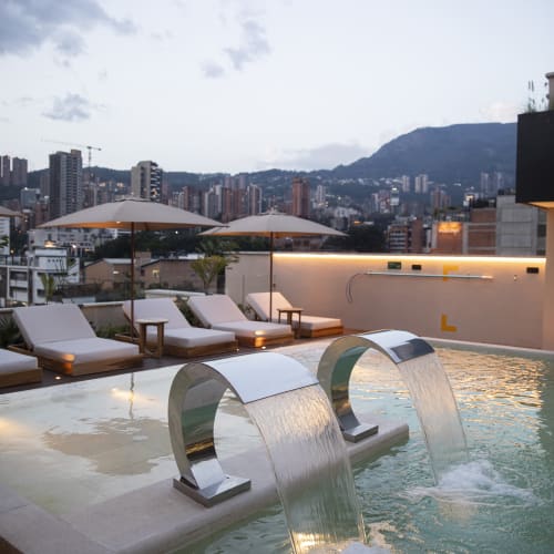 Moderner Dachpool mit zwei gebogenen Wasserfontänen, Liegestühlen und Sonnenschirmen mit Blick auf eine Stadtlandschaft mit Bergen im Hintergrund bei Dämmerung.