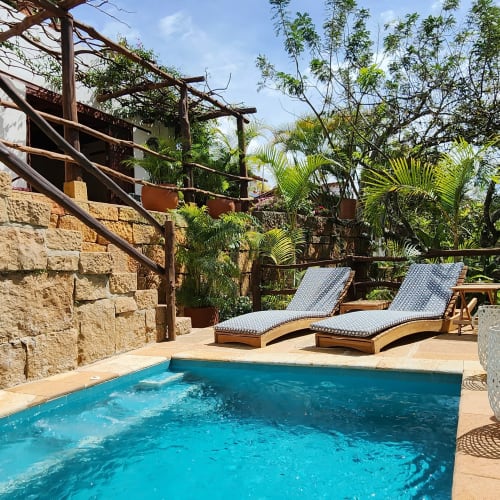 Small rectangular outdoor pool with two lounge chairs on the deck surrounded by stone walls and lush greenery under blue sky.