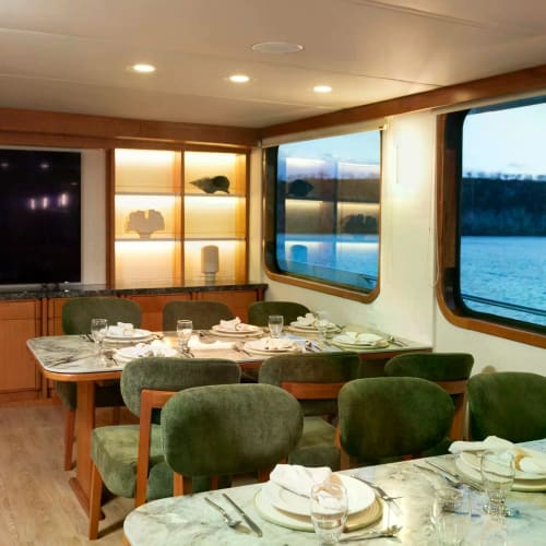 Dining area inside a yacht with marble-topped tables set with plates, glasses, and silverware, surrounded by green upholstered chairs, and large windows showing a water view outside.