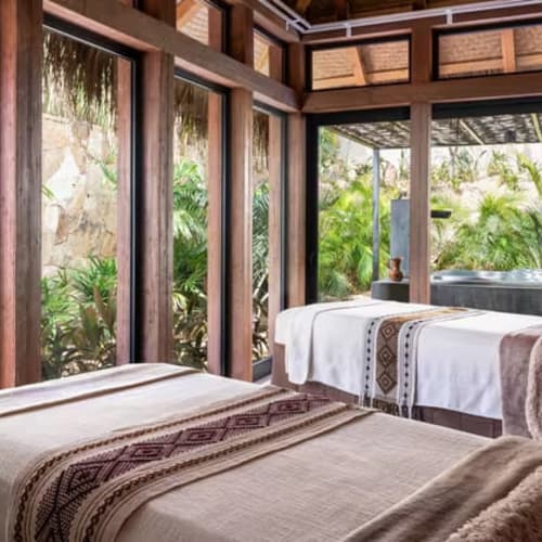 Zwei Massageliegen in einem Spa-Raum mit großen Fenstern, die auf üppiges Grün und einen Whirlpool im Freien blicken, mit natürlichen Holzbalken und sanfter Beleuchtung.