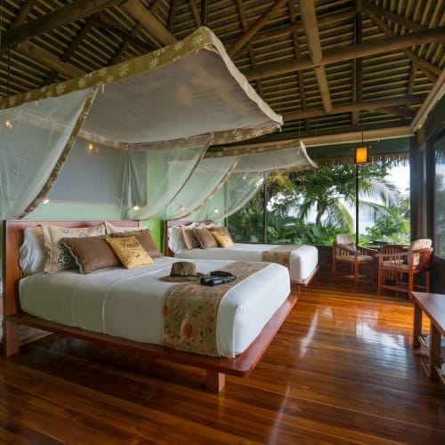 Luxurious bungalow bedroom with twin beds, mosquito net canopy, and large windows overlooking greenery and the ocean in Costa Rica.