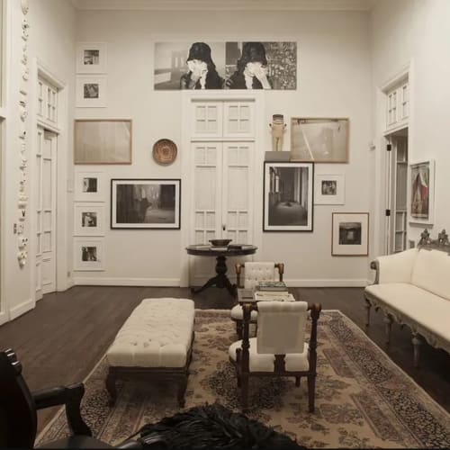 Innenraum eines elegant eingerichteten Galerieraums mit Vintage-Möbeln, darunter weiße gepolsterte Stühle, ein Sofa, ein Holztisch und gerahmte Schwarz-Weiß-Fotos an den Wänden.