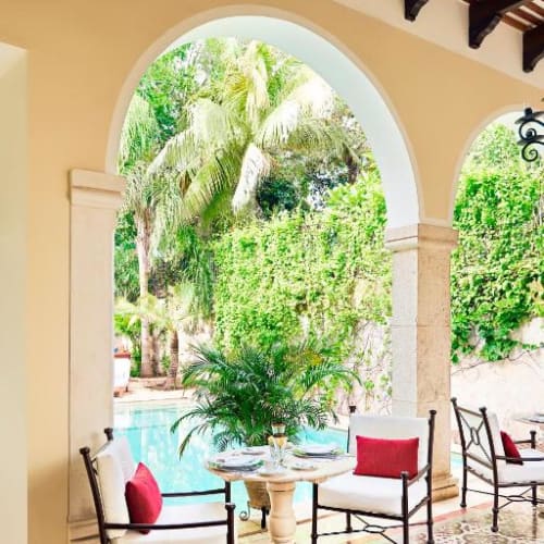 Überdachte Terrasse mit gewölbten Bögen und schmiedeeisernen Stühlen mit weißen Polstern und roten Kissen um einen kleinen runden Tisch, mit Blick auf einen Pool und üppiges Grün mit Palmen im Hintergrund.