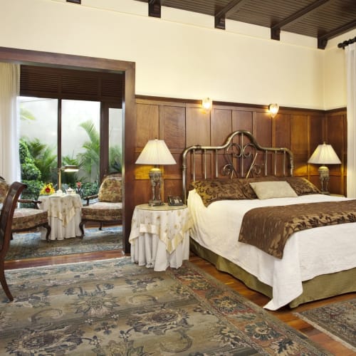 Ein gemütliches Hotelzimmer mit großem Bett, zwei Nachttischlampen und einem Sitzbereich mit Tisch und Stühlen in der Nähe eines Gartens.
