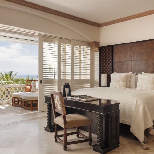 Ein luxuriöses Schlafzimmer mit einem großen Bett, versehen mit cremefarbenen Kissen und einer strukturierten Decke, einem dunklen Holztisch mit Stuhl und einer geöffneten Tür, die zu einer Terrasse mit Meerblick und Sitzgelegenheiten im Freien führt.