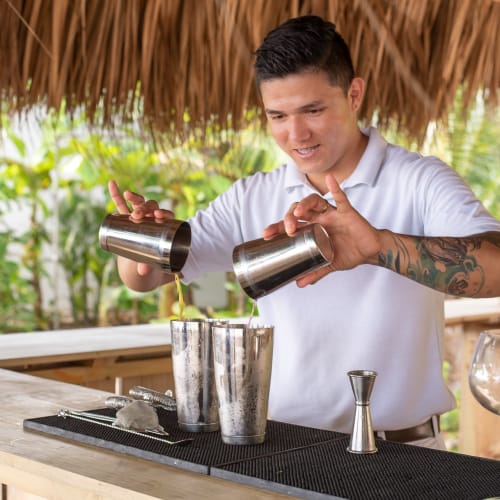 A bartender pouring drinks into metal cocktail shakers at an outdoor beach bar with a thatched roof.