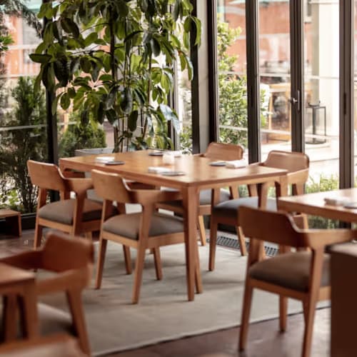 Holztische und -stühle in einem Restaurant mit großen Glasfenstern und grünen Pflanzen draußen.