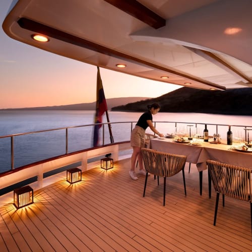 Eine Person deckt einen Esstisch auf dem Deck einer Yacht bei Sonnenuntergang mit Blick auf das Meer und entfernte Hügel.