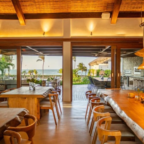 Innenraum eines warm beleuchteten Restaurants mit Holztischen und Stühlen, große Glastüren öffnen sich zu einer Außenterrasse mit Palmen und Meerblick.