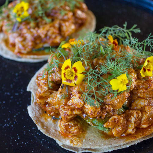 Zwei Portionen Garnelen-Tapas, garniert mit frischem Dill und essbaren gelben Blüten auf einem schwarzen Teller.