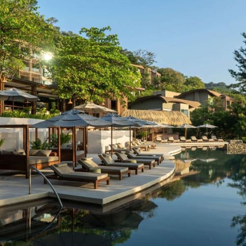 Luxusresort-Pool mit Sonnenliegen und Sonnenschirmen, umgeben von grünen Bäumen und Gebäuden am Hügel.