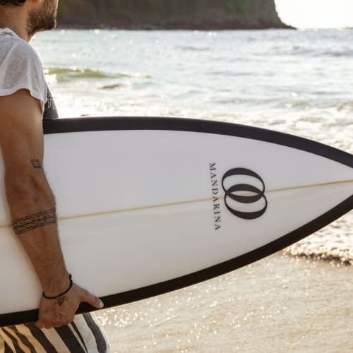 Ein Mann hält ein weißes Mandarina-Surfbrett mit schwarzen Kanten und steht am sandigen Strand nahe dem Ozean.