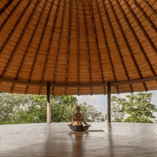 Eine Frau praktiziert Yoga in einem großen, offenen Holzpavillon mit grünem Umfeld unter einem hohen, runden Strohdach.