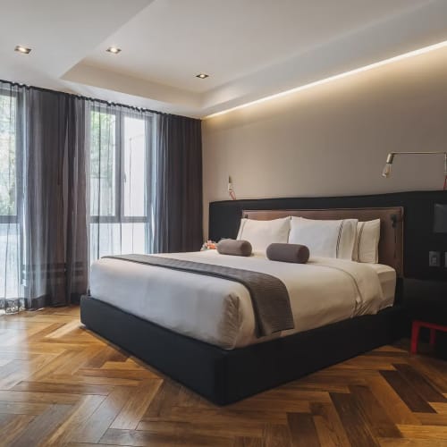 Modernes Executive-Schlafzimmer mit großem Bett, weißer Bettwäsche und zwei lila Nackenrollen, neben Fenstern mit transparenten und dunklen Vorhängen sowie warmem Holzparkettboden.