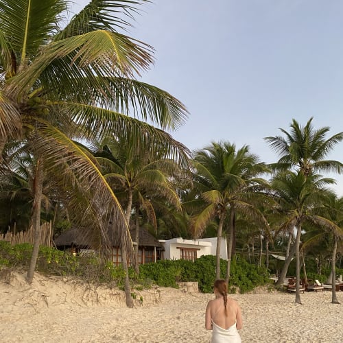 Eine Frau in einem weißen Kleid läuft am Sandstrand entlang, gesäumt von hohen Palmen und tropischem Grün, im Hintergrund sind rustikale Hütten und ein modernes weißes Gebäude zu sehen.