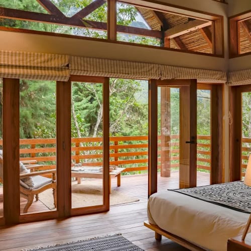 Ein gemütliches Schlafzimmer mit großen Glasschiebefenstern, die auf einen Holzbalkon mit üppigem grünen Baumbewuchs führen.