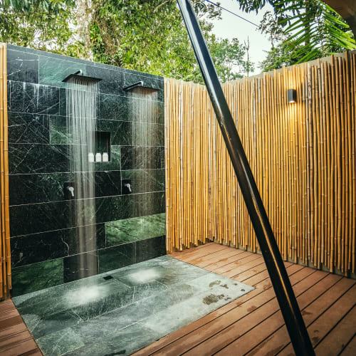 Outdoor shower area with two rainfall showerheads mounted on a black tiled wall, surrounded by a bamboo fence and wooden floor in a tropical setting.