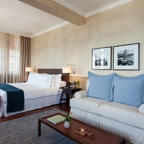A modern hotel room featuring a double bed with white linens and blue throw, a beige sofa with large blue cushions, and a wooden coffee table with a book and decorative items, set against beige walls with two framed black and white photographs.