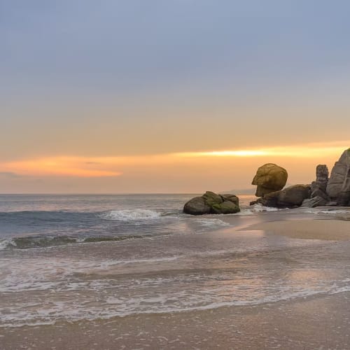 Ruhiger Strand bei Sonnenuntergang mit sanften Wellen, die am Ufer ankommen, und großen Felsen an einer Klippe rechts.